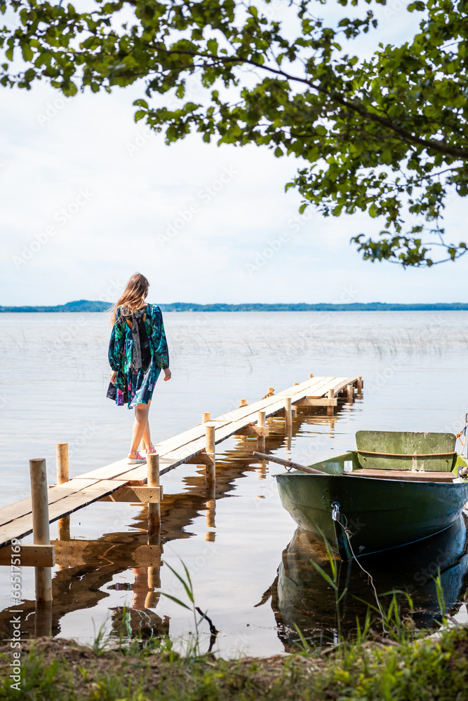 Fototapeta premium A woman in a dress in sunset light walks along a wooden bridge on the shore of the lake