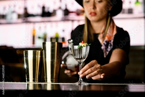 Young bartender girl in black clothes holding clean empty glass on long leg at bar counter