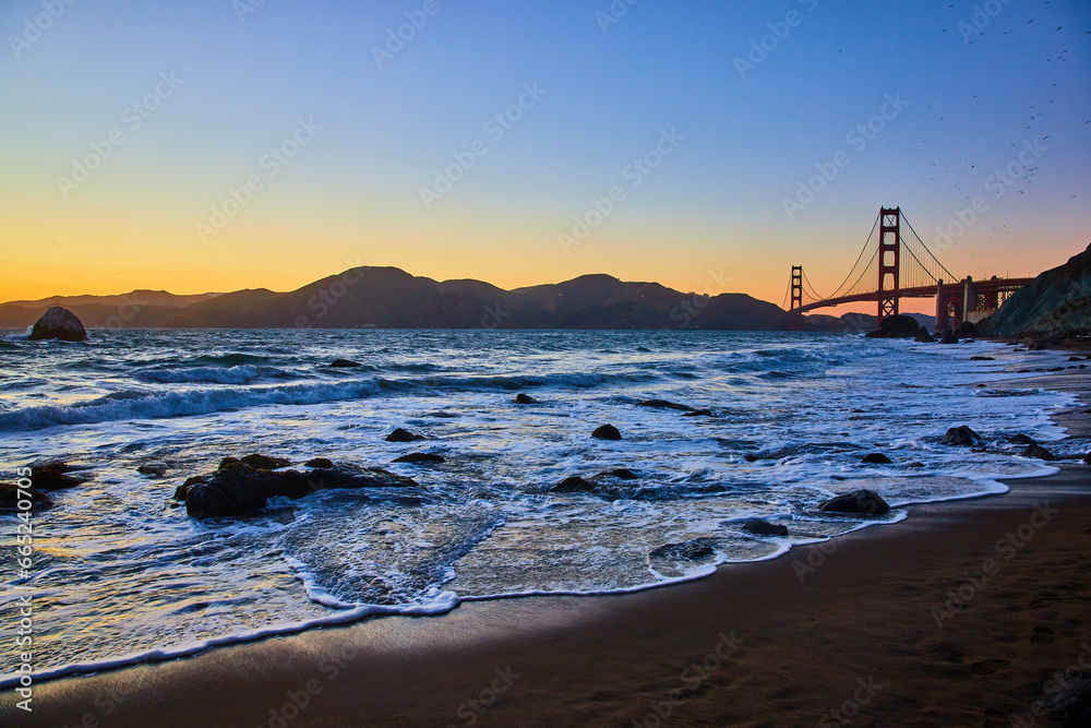 Glowing blue waves with sunset sky and Golden Gate Bridge along sandy shore