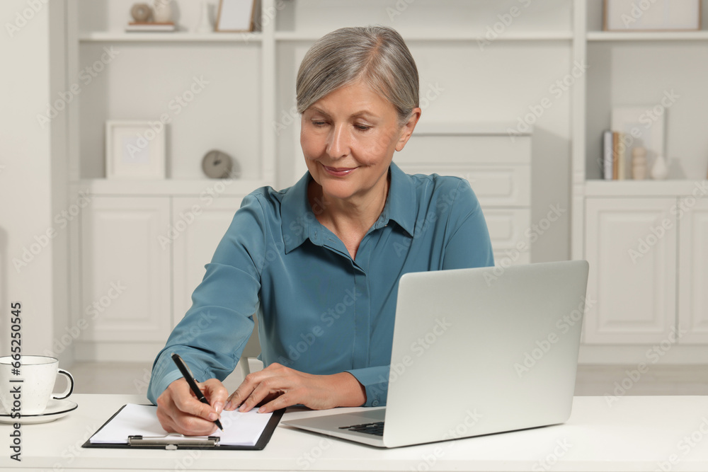 Beautiful senior woman taking notes near laptop at white table indoors Beautiful senior woman taking notes near laptop at white table indoors