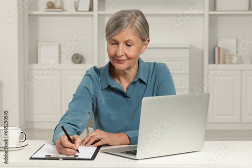 Beautiful senior woman taking notes near laptop at white table indoors