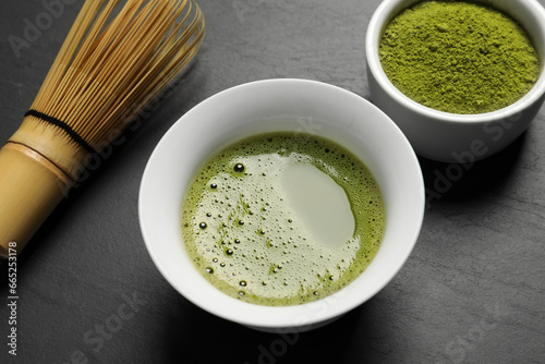Cup of fresh matcha tea, green powder and bamboo whisk on black table, closeup