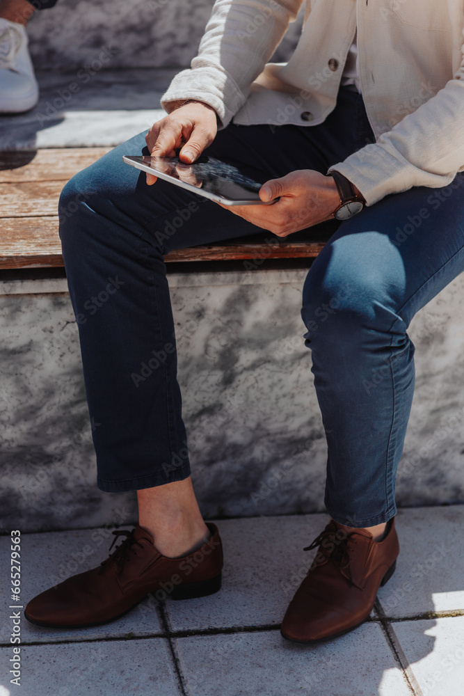 Fototapeta premium Stylish businessman sitting on a bench outdoors holding tablet. Cropped photo
