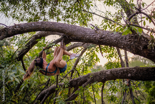 An active and fit girl exercises outdoors, climbing a tree in a green park. She is strong, flexible, and enjoys a healthy lifestyle while having fun in a beautiful natural environment.