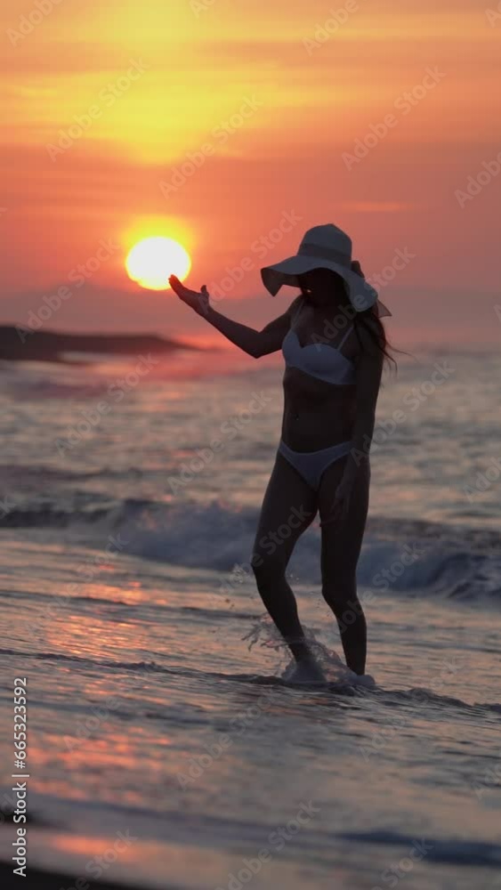 Full length woman in white bikini and straw sun hat on beach at sunrise. Female walking ankle deep in water in breaking waves during summer beach holiday. Slow motion, handheld shot, vertical video Full length woman in white bikini and straw sun hat on beach at sunrise. Female walking ankle deep in water in breaking waves during summer beach holiday. Slow motion, handheld shot, vertical video