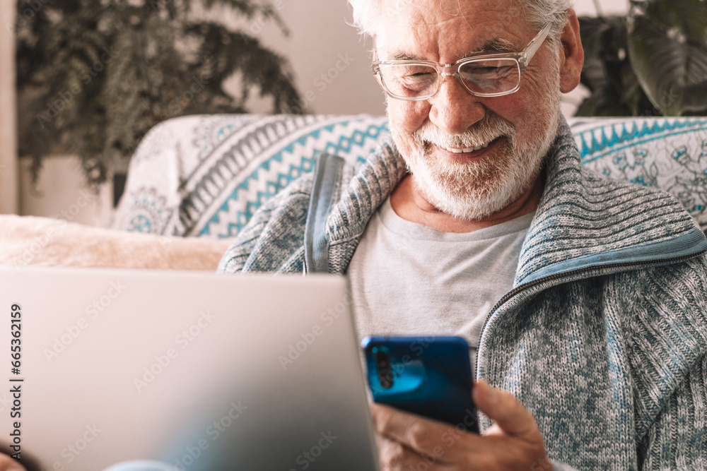 Smiling senior caucasian man using laptop and smart phone sitting on couch at home. Modern retired bearded male enjoying tech and social news. Retirement leisure concept Smiling senior caucasian man using laptop and smart phone sitting on couch at home. Modern retired bearded male enjoying tech and social news. Retirement leisure concept