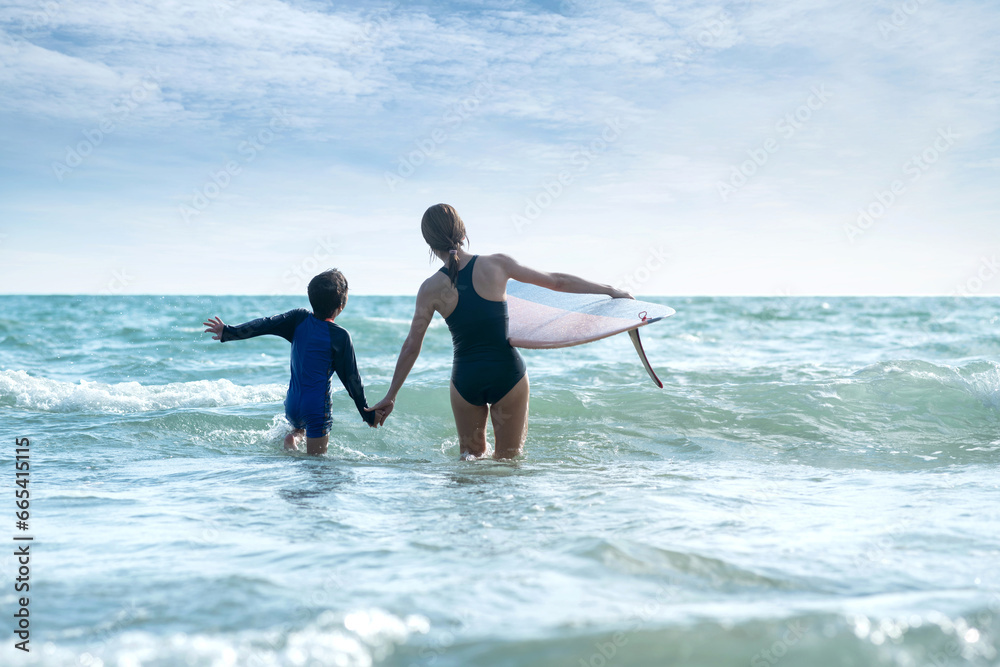 Back view of of happy mother and child son walking hand in hand in the sea, mother holding a surfboard with the other hand, lifestyle activities, water sports