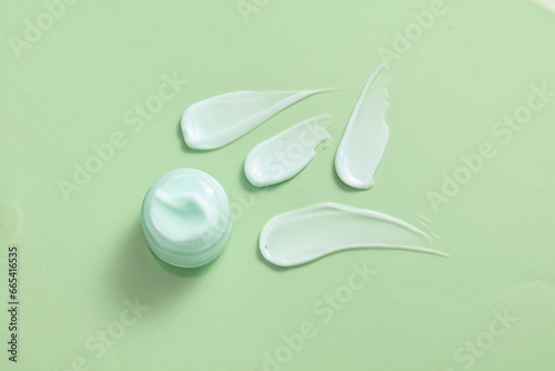 An unlabeled jar of cosmetic filled with a dense texture, surrounded by four streaks of moisturizer on a light green background. The moisturizer provides essential active element for the skin. photo