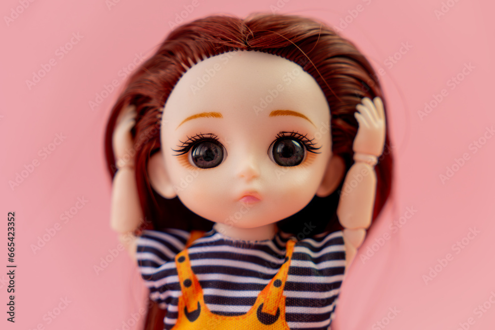 child toy doll with dark hair in an orange dress standing on a pink background. Plastic children's toy. Doll games for imagination. the frightened doll holding his head in fear. child toy doll with dark hair in an orange dress standing on a pink background. Plastic children's toy. Doll games for imagination. the frightened doll holding his head in fear.