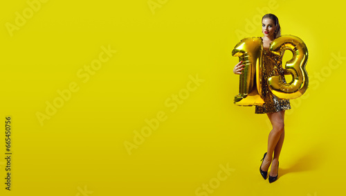 A slender young tanned woman in a shiny dress holds the number 13 folded from golden inflatable balloons. Holiday, birthday. Number 13. Model posing on a golden background. 