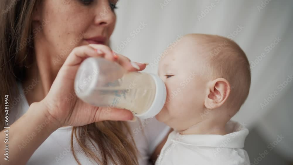 Mother feeding baby from bottle on kitchen at home stroking kid on head. Tiny infant sitting in baby chair. Little child eating milk mix. Baby care, maternity leave, babyhood, motherhood care concept. Mother feeding baby from bottle on kitchen at home stroking kid on head. Tiny infant sitting in baby chair. Little child eating milk mix. Baby care, maternity leave, babyhood, motherhood care concept.