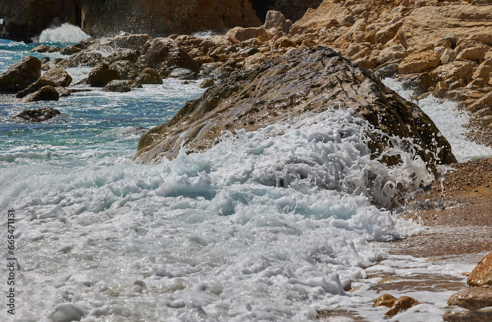 Waves waves crashing against rocks on the shores of the Mediterranean Sea during the day. Cala del Moraig beach in Benitachell of Alicante, Spain.