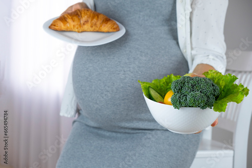 Belly of pregnant woman with bowl of vegetables and fruits and croissant. Craving of sweets and dessert during pregnancy. Diet while gestational diabetes. photo