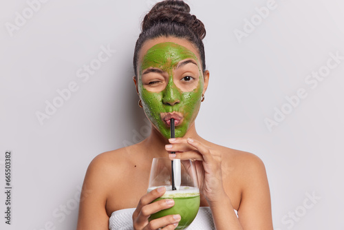 Horizontal shot of dark haired Latin woman drinks fresh smoothie applies nourishing green facial mask made on natural products wrapped in bath towel isolated over white bacground. Detox concept