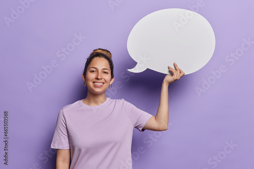 Waist up shot of cheerful young woman holds white communication bubble with area for promotional content suggests to write your ideas here dressed in t shirt isolated over purple background.