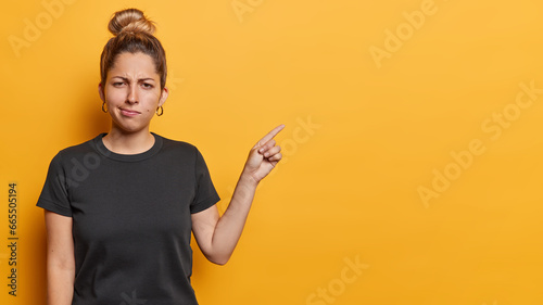 Fotografia Waist up shot of discontent sulking woman frowns face indicates at blank copy space with frustrated expression dressed in casual black t shirt isolated on yellow background