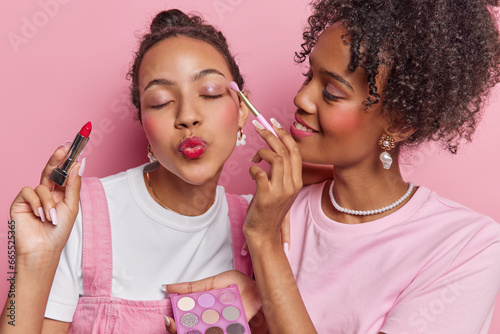 Photo of curly haired woman applies eyeshadow to friend do makeup together put on lipstick prepare for dating want to have fabulous look stand closely to each other isolated over pink background