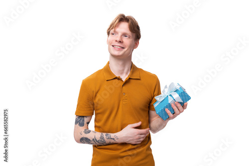 young handsome red-haired guy dressed in an orange t-shirt with a tattoo on his arm holds a gift box for valentine's day