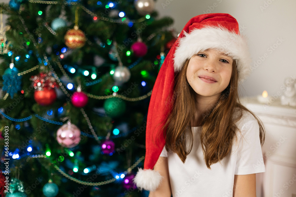 Close-up portrait of a smiling teenage girl wearing a Santa hat on the background of a Christmas tree