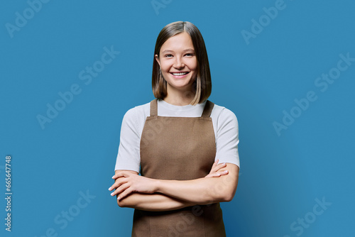Portrait of young smiling confident woman in apron on blue background