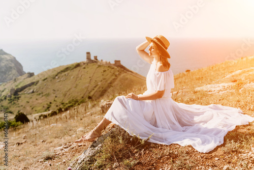 Happy woman in a white dress and hat stands on a rocky cliff above the sea, with the beautiful silhouette of hills in thick fog in the background.