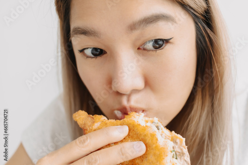 Close up of asian woman holding and having homemade breakfast, eating sandwich hungrily at home alone while looking at something.