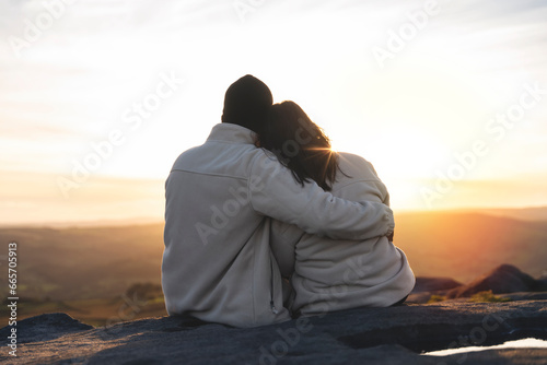 Fotografie Back view of the happy couple in love sitting on top of a mountain enjoying a su