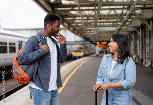 Happy loving couple with luggage and backpack walking along railway platform as they are ready to travel on holiday. Man and woman holding  hands,  looking back and smiling. Travel by train concept