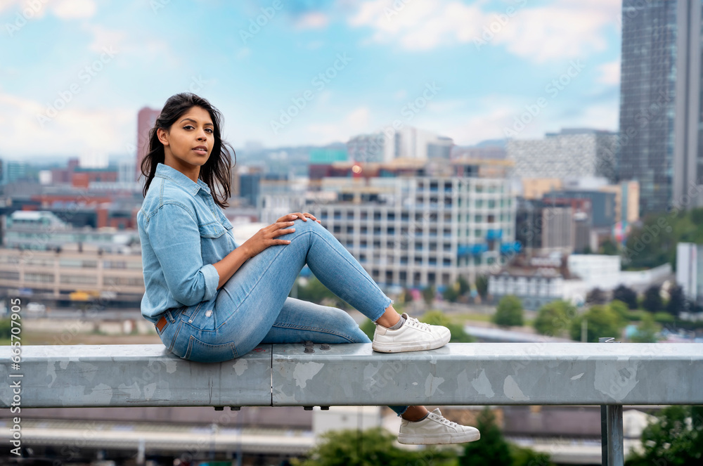 Portrait of a beautiful  Indian woman sitting  on the barrier in front of a modern city background dreaming about the future