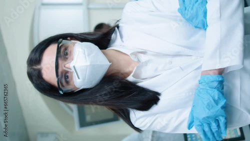 Vertical orientation of beautiful female pharmacist or biologist with respirator and goggles looking seriously at camera. Smart woman wearing gloves and crossing hands while standing in laboratory. photo