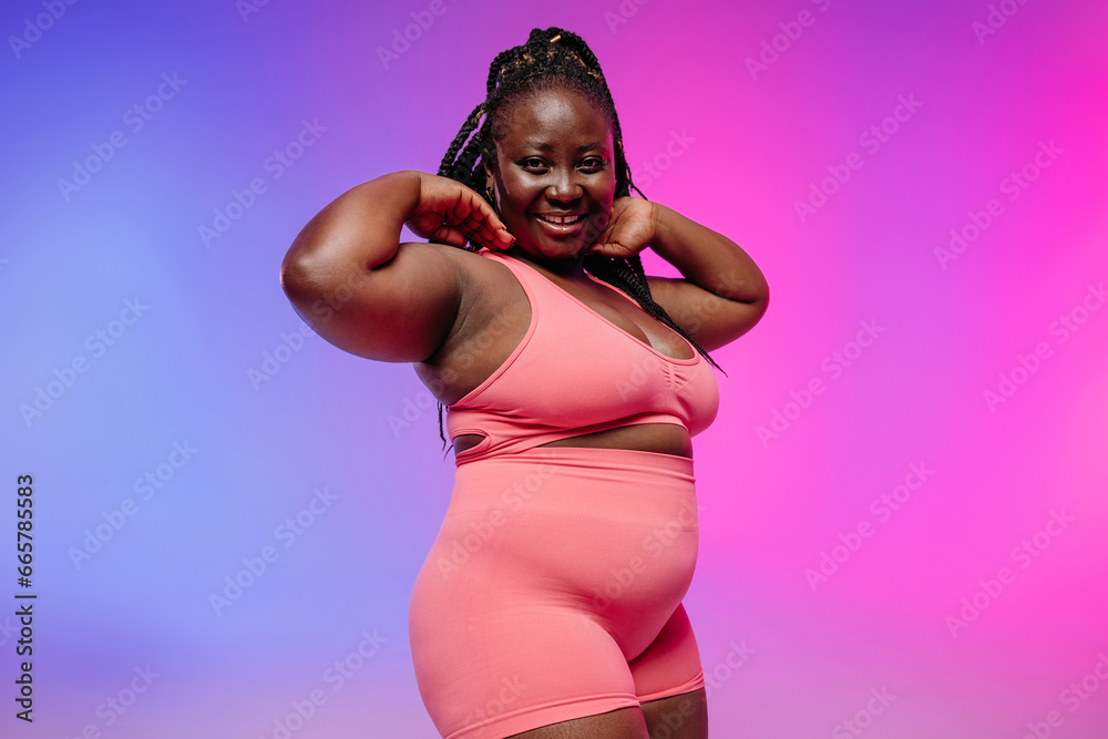 Obraz premium Voluptuous African woman in sportswear radiating self-love while standing on vibrant background