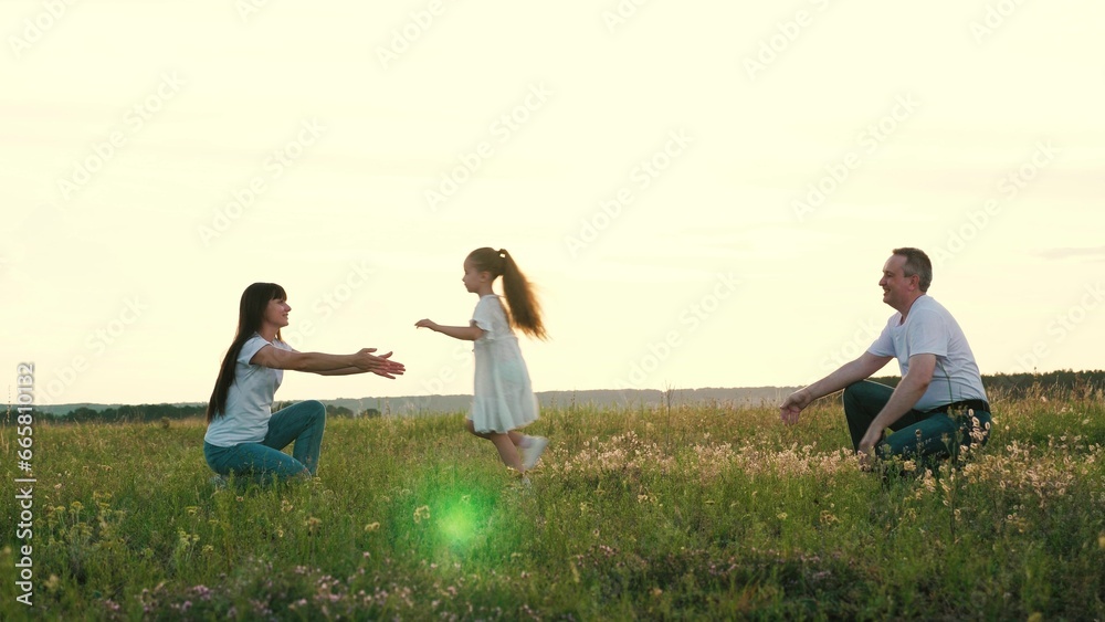 Happy little girl runs between mother and father to hug parent resting in summer field