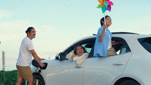 Concept of progressive happy family enjoying their time at wind farm with electric vehicle. Electric vehicle driven by clean renewable energy from wind turbine generator for charging station. © Summit Art Creations
