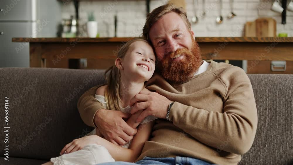 Smiling bearded man hugging daughter and looking at camera on couch at home Smiling bearded man hugging daughter and looking at camera on couch at home