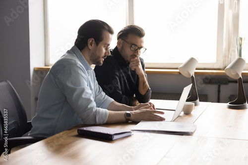 Young serious businesspeople discuss new software use notebook, engaged teamwork at workplace. Successful entrepreneur consulting client in office, show new presentation. Business, meeting, brainstorm