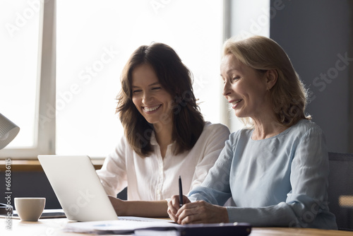 Middle aged woman mentor help assisting to new employee at workplace, prepare presentation, do work task. Cheerful colleagues looking at laptop screen work together in office. Teamwork, partnership