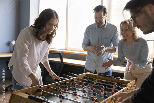 Four positive diverse older and younger employees engaged in foosball game in modern office, play together tabletop soccer indoors, spend carefree break at workplace. Hobby, teambuilding activity, fun photo