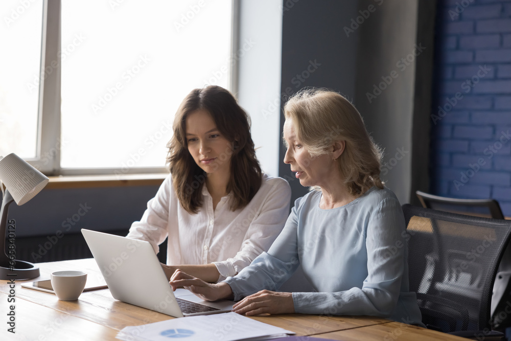 Younger 35s and older 60s women colleagues having common task work together sit at desk use laptop, talk, discuss on-line project, learn software or corporate program. Teamwork, tech, apprenticeship Younger 35s and older 60s women colleagues having common task work together sit at desk use laptop, talk, discuss on-line project, learn software or corporate program. Teamwork, tech, apprenticeship