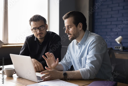 Two young focused business partners sit at desk in modern office discuss project details, share ideas, mentor teaches employee new business application, explains online work. Collaboration, teamwork