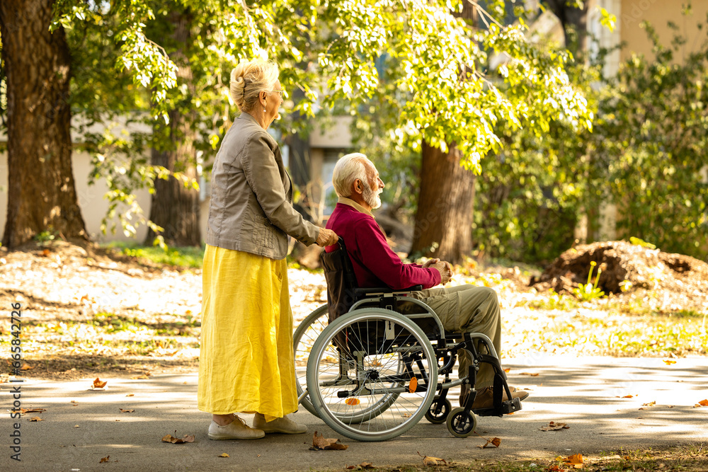 Elderly woman pushing wheelchair with senior man in nursery home park. Elderly woman pushing wheelchair with senior man in nursery home park.