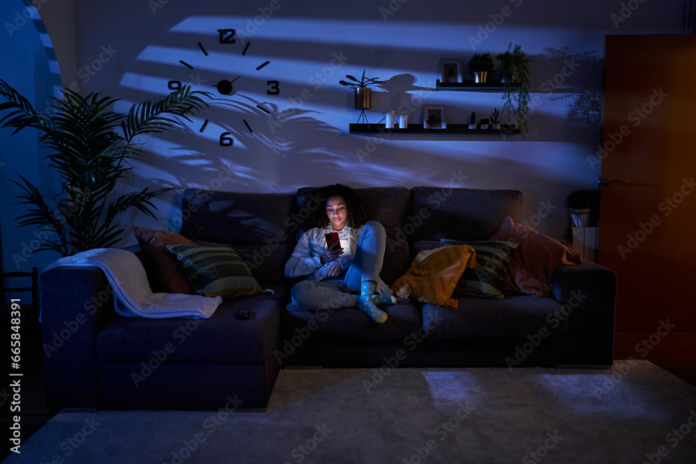 Naklejka premium Young woman with serious expression looking at cell phone sitting in living room at night. Concentrated latina female using mobile on sofa at home. Copy space, generation z and free time indoors.
