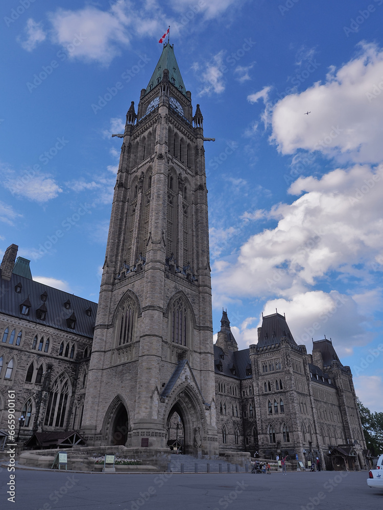 Captured from a distance, the panoramic view of Centre Block of the Canadian Parliament Building reveals its grandeur, set against a vast backdrop of azure skies and billowing white clouds.