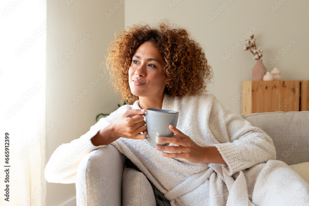 Pensive multiracial woman relaxing at home, sitting on the sofa drinking tea. - obrazy, fototapety, plakaty 