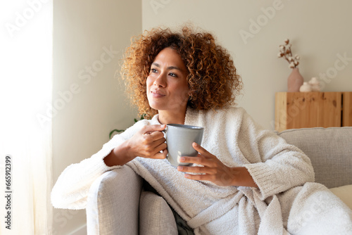 Pensive multiracial woman relaxing at home, sitting on the sofa drinking tea.