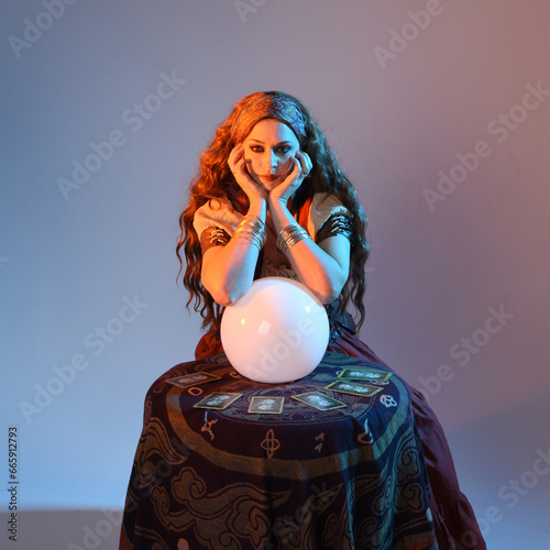 Photography close up portrait of beautiful red haired woman wearing a medieval fantasy fortune teller costume, looking into crystal ball reading the future at seance table