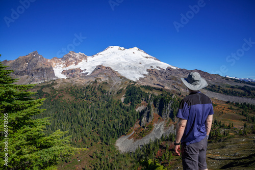 Adventurous athletic male hiker standing on top of a hill looking at Mount Baker on a beautiful sunny fall day in the Pacific Northwest.
