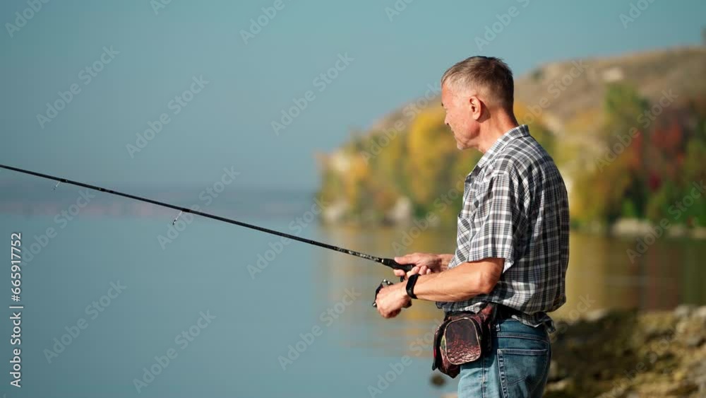 An adult man in casual clothes catches fish with a spinning rod. A mature man spends time on the river bank fishing on a warm sunny day. He turns the reel intently, hoping for a catch. An adult man in casual clothes catches fish with a spinning rod. A mature man spends time on the river bank fishing on a warm sunny day. He turns the reel intently, hoping for a catch.