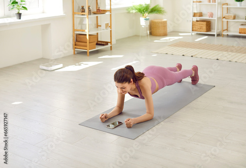 Portrait of a young sporty woman doing plank sport exercise lying on yoga mat on the floor in the living room at home and looking on mobile phone screen. Fitness, fit and home training concept.