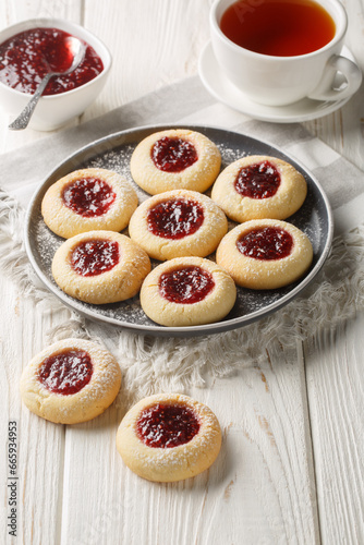 Thumbprint Christmas cookies filled with raspberry jam closeup on the plate served with tea on the white wooden table. Vertical photo