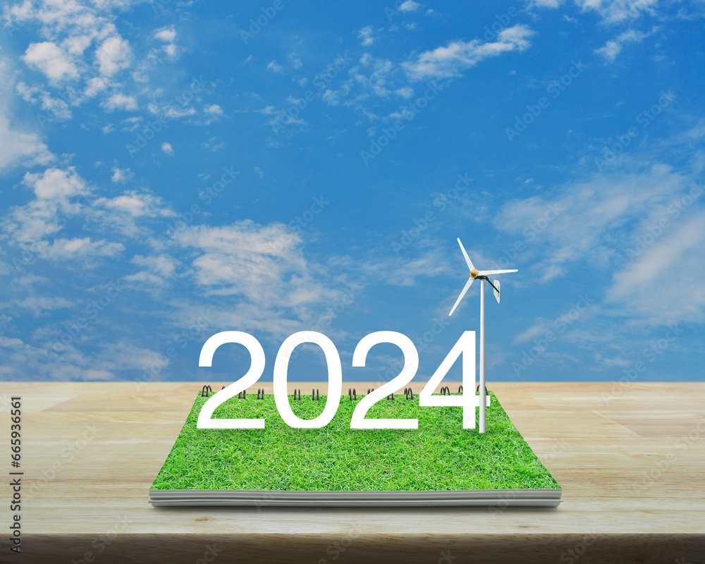 Obraz premium 2024 white text with wind turbine on green grass on open book on wooden table over blue sky with white clouds, Happy new year 2024 ecological cover concept